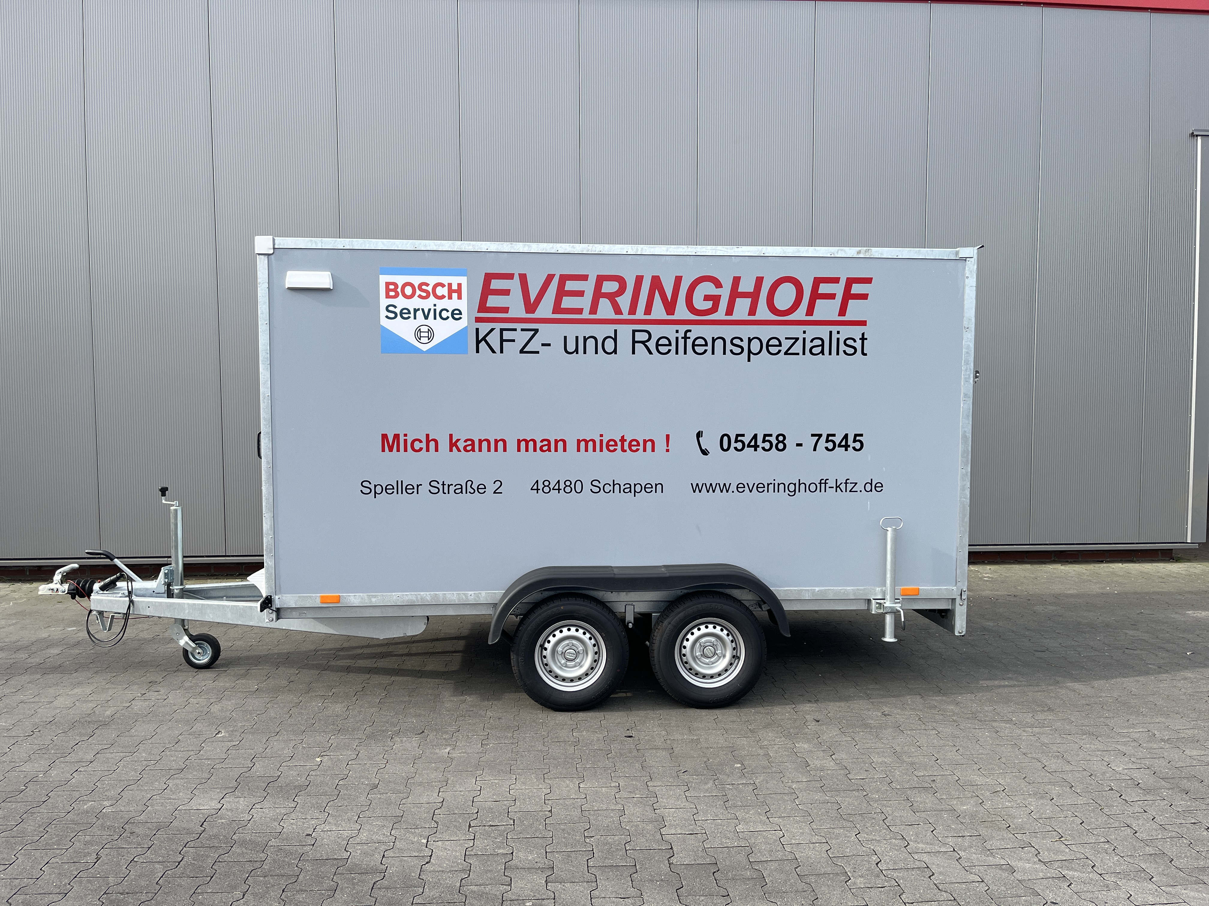 Weisser Kofferanhaenger mit Werbung der Firma Everinghoff vor einer grauen Wand